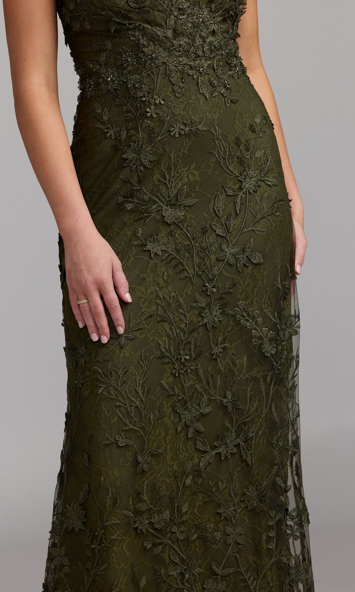 A person wearing the Long Beaded Formal Dress: Mac Duggal 68192—an elegant olive green gown adorned with intricate beading and floral embroidery—poses with one hand resting by their side and the other near their hip, set against a plain gray background.